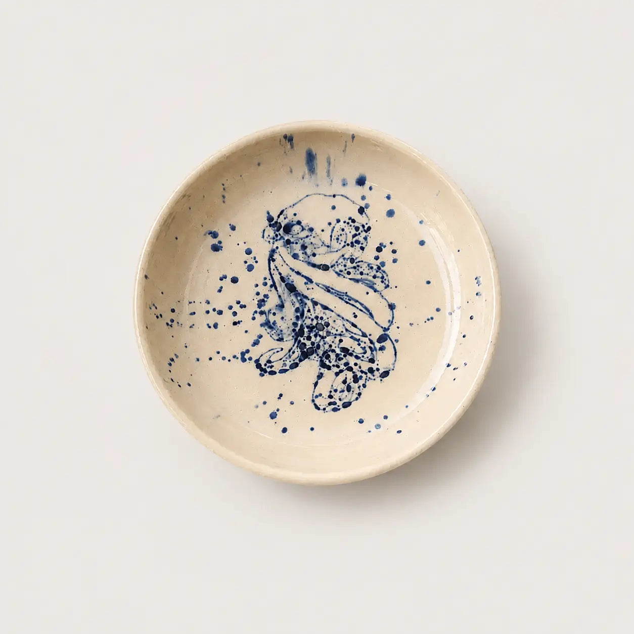 A round ceramic small plate with a speckled beige glaze and a hand-painted blue floral design in the center, photographed from directly above on a light beige background.