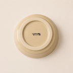 The underside of a round ceramic small plate or dish in a warm cream-beige glaze, featuring the black printed brand name 'CARDON STUDIO' in the center, set against a matching light beige background.