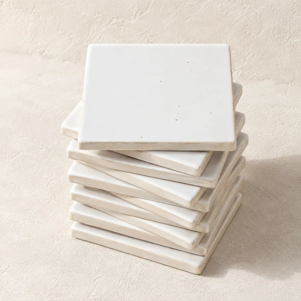 A stack of square white ceramic coasters with a matte finish, arranged on a textured beige surface against a matching beige background.