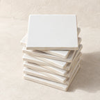 A stack of square white ceramic coasters with a matte finish, arranged on a textured beige surface against a matching beige background.
