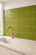 A modern kitchen backsplash featuring glossy olive green square tiles with a woodgrain texture, installed behind a light wood countertop and a copper-colored faucet.