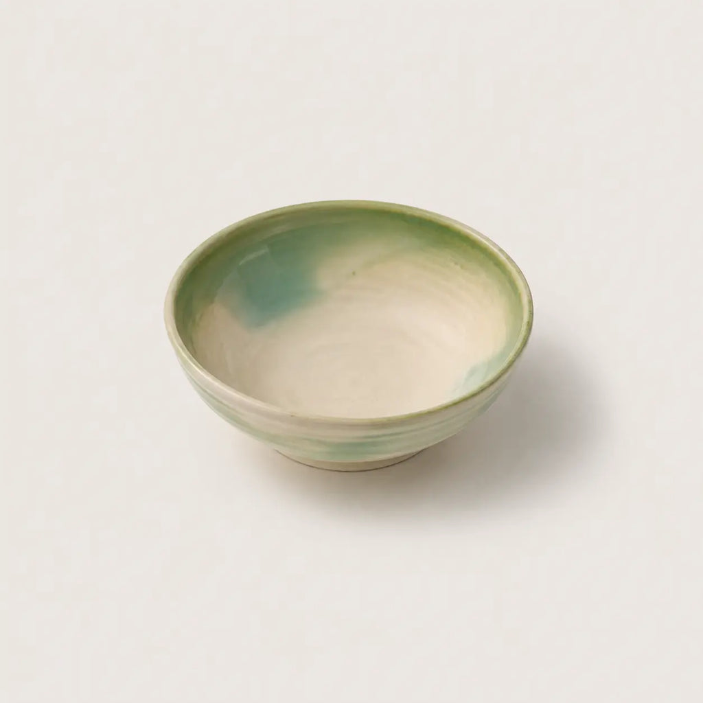 A small handmade ceramic snack bowl with a glossy finish, featuring a light cream interior with soft green and teal glaze accents, set against a plain off-white background.
