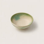 A small handmade ceramic snack bowl with a glossy finish, featuring a light cream interior with soft green and teal glaze accents, set against a plain off-white background.