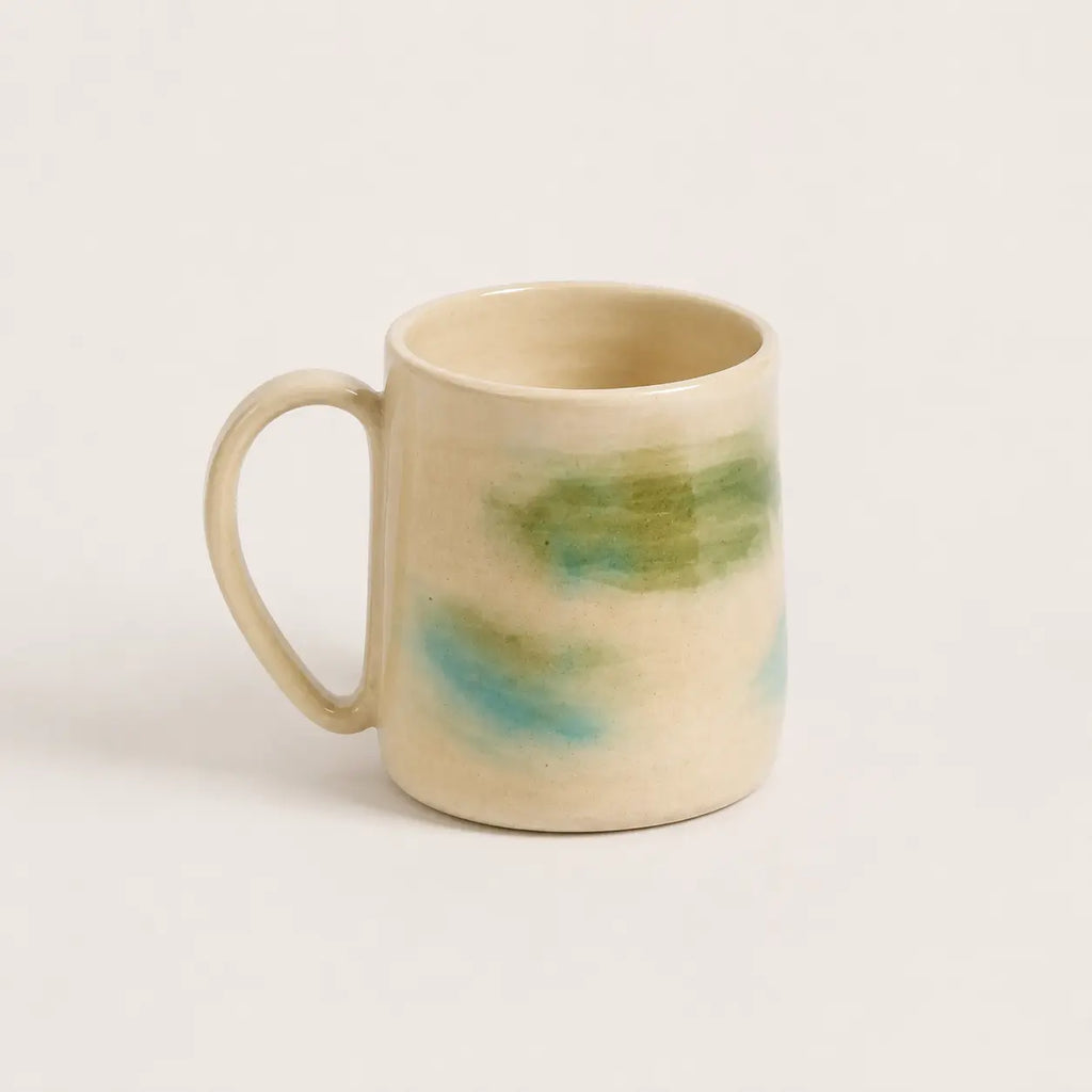 A handmade porcelain mug with a warm cream-beige glaze and abstract painted accents of green and light blue, shown on a plain light background.
