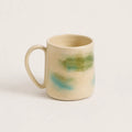 A handmade porcelain mug with a warm cream-beige glaze and abstract painted accents of green and light blue, shown on a plain light background.