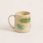 A handmade porcelain mug with a warm cream-beige glaze and abstract painted accents of green and light blue, shown on a plain light background.