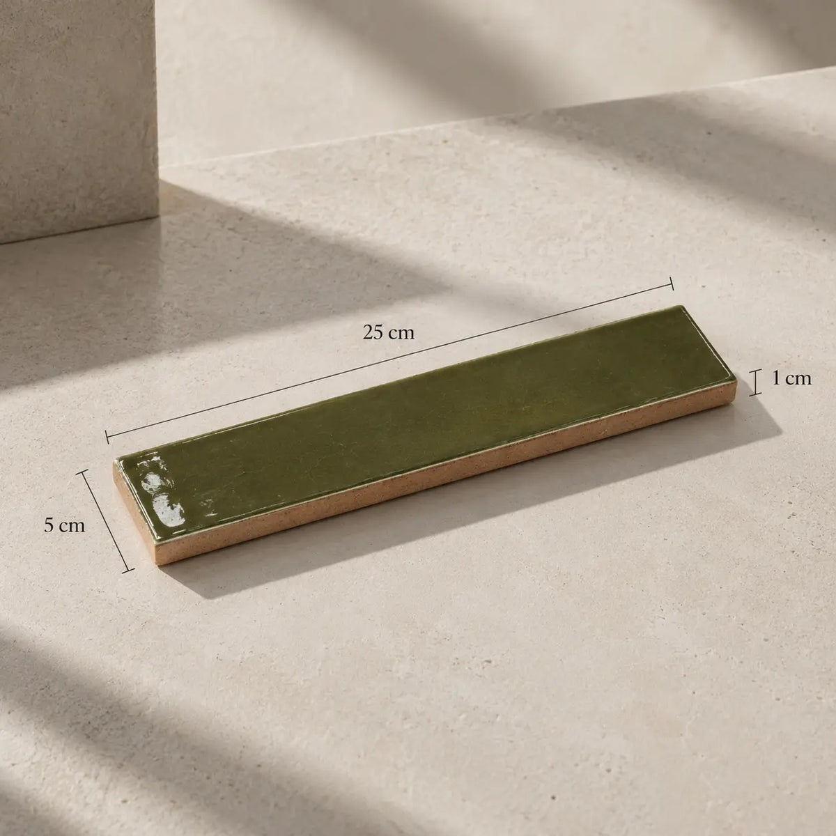 A rectangular ceramic tile with a glossy olive green top and a natural beige clay edge, shown on a light beige stone surface with dimension lines indicating 25 cm length, 5 cm height, and 1 cm thickness.