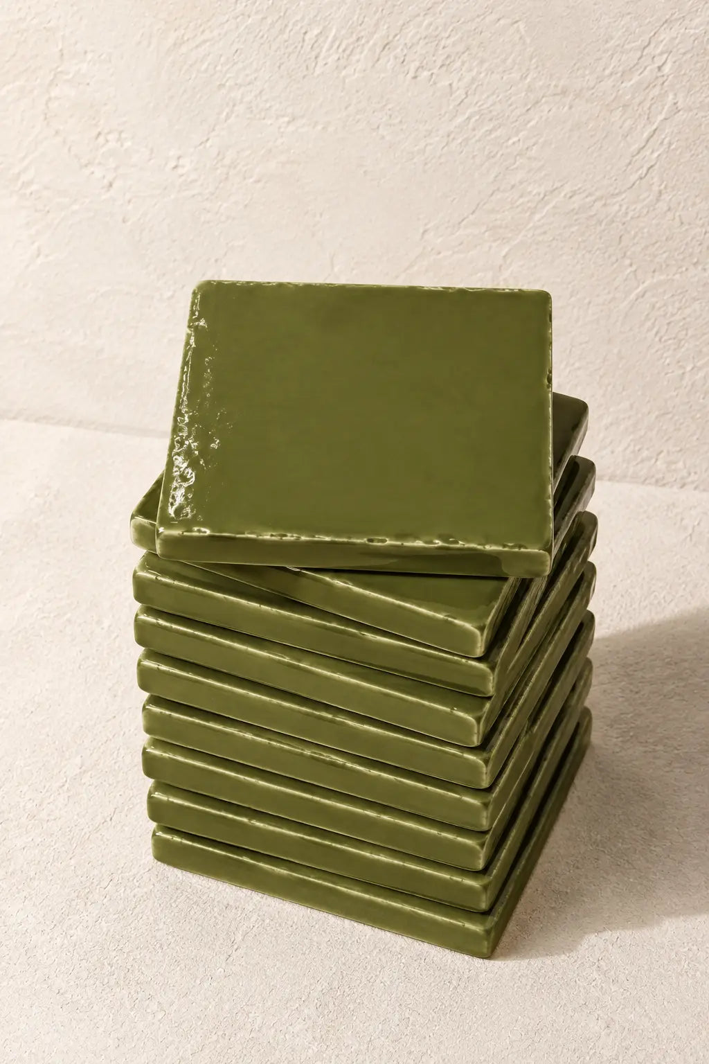 A stack of square olive green coasters with a glossy finish, arranged on a textured off-white surface against a matching background.