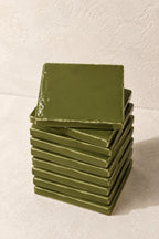 A stack of square olive green coasters with a glossy finish, arranged on a textured off-white surface against a matching background.