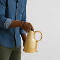 A man in a blue denim shirt holds a light beige ceramic pitcher with a loop handle against a plain light gray background.