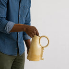 A man in a blue denim shirt holds a light beige ceramic pitcher with a loop handle against a plain light gray background.