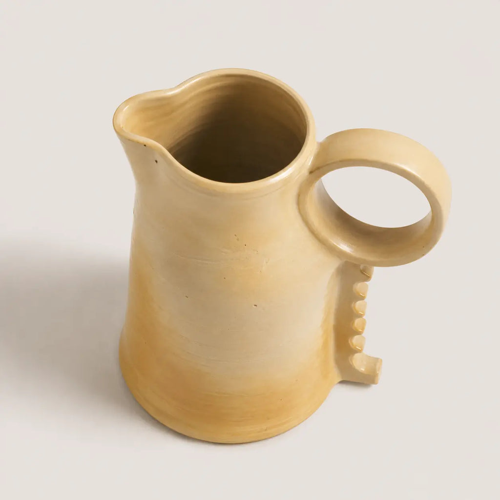 A handmade ceramic pitcher with a warm beige to light brown gradient glaze, featuring a loop handle and a small decorative tab on the side, set against a plain light gray background.