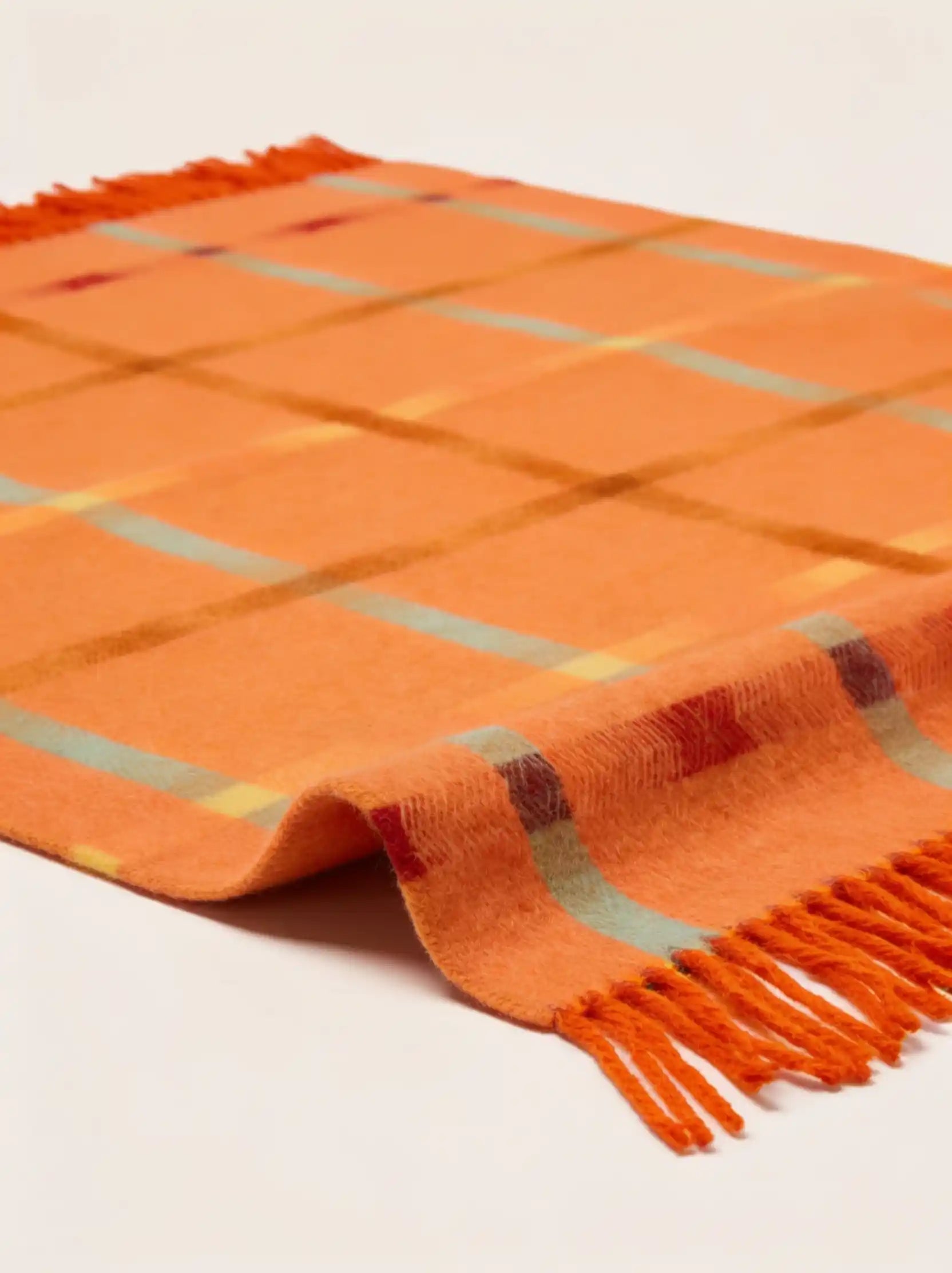 An orange plaid throw blanket with fringe ends, featuring intersecting stripes of yellow, light blue, and dark red, shown on a clean off-white background.