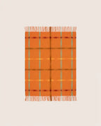 An orange woven throw blanket with a plaid pattern in muted green, yellow, and burgundy, featuring fringed edges on a light cream background.
