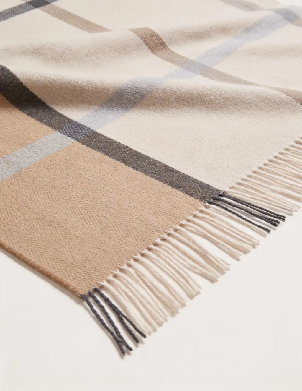 A close-up of a woven plaid throw blanket throw or throw blanket with cream, beige, light gray, and dark charcoal stripes, finished with fringed edges.