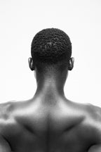 Black and white studio photograph of a man's bare back and neck, viewed from behind, with short curly hair and no visible clothing against a plain light background.