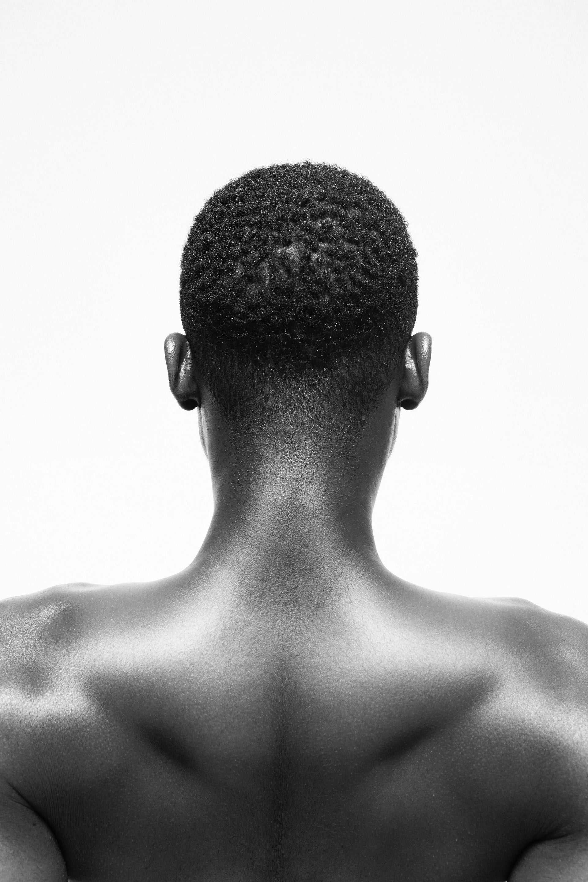 Black and white studio photograph of a man's bare back and neck, viewed from behind, with short curly hair and no visible clothing against a plain light background.