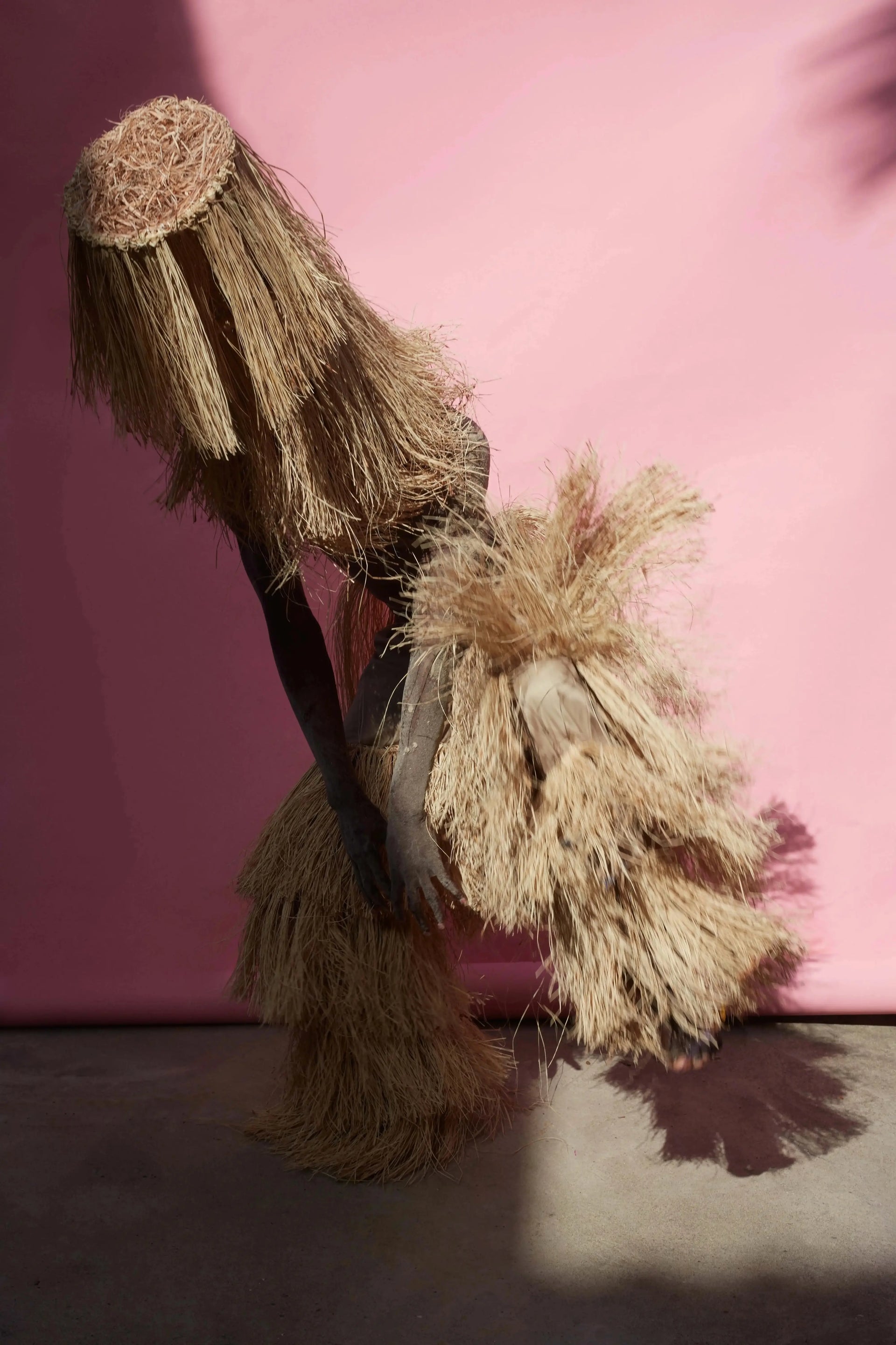 A person wearing a voluminous natural-fiber costume or dress with a matching fringed headpiece, posed against a pink wall on a concrete floor.