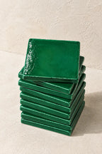 A stack of glossy green square coasters on a textured beige surface against a matching beige wall.