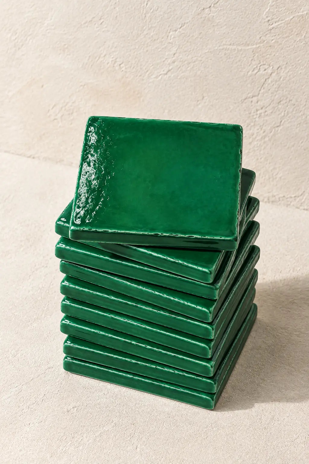 A stack of glossy green square coasters on a textured beige surface against a matching beige wall.