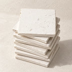 A stack of square off-white stone coasters with a textured, rustic finish, photographed on a matching light beige surface.