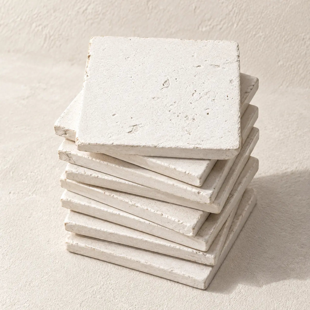 A stack of square off-white stone coasters with a textured, rustic finish, photographed on a matching light beige surface.
