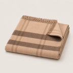 A folded beige plaid throw blanket with fringed edges, displayed on a clean off-white background.