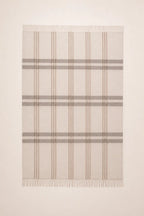 A rectangular woven throw blanket with fringed edges, featuring a classic plaid pattern in beige, taupe, and light brown on a cream background.