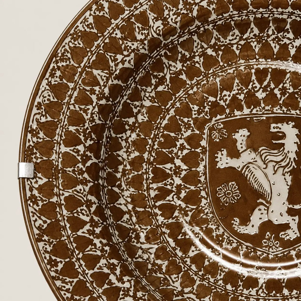 A close-up of a decorative ceramic plate featuring a brown and white hand-painted pattern with a heraldic lion crest in the center.