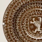 A close-up of a decorative ceramic plate featuring a brown and white hand-painted pattern with a heraldic lion crest in the center.