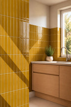 Bright kitchen corner with glossy amber yellow wall tiles, light wood cabinetry, a white countertop, and a window with natural light.
