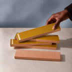 Hand holding a glossy amber yellow rectangular ceramic tile above a stack of handmade tiles on a neutral stone surface.
