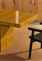 Glossy amber yellow rectangular ceramic tiles covering a dining table, paired with a dark wood chair and natural woven rug.