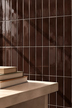 Glossy dark brown rectangular ceramic tiles arranged in a grid pattern on a wall, with a light beige stone ledge in the foreground holding a stack of books.
