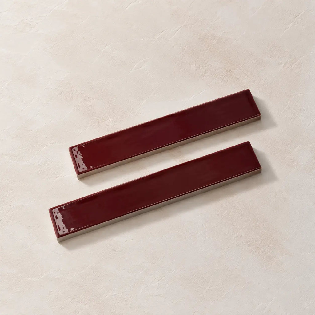 Two glossy rectangular cabinet pulls in a deep burgundy red finish, displayed diagonally on a textured beige surface.