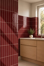 A modern kitchen corner featuring glossy deep red ceramic tiles on the wall, light wood cabinetry, a white countertop, and a window with natural light.
