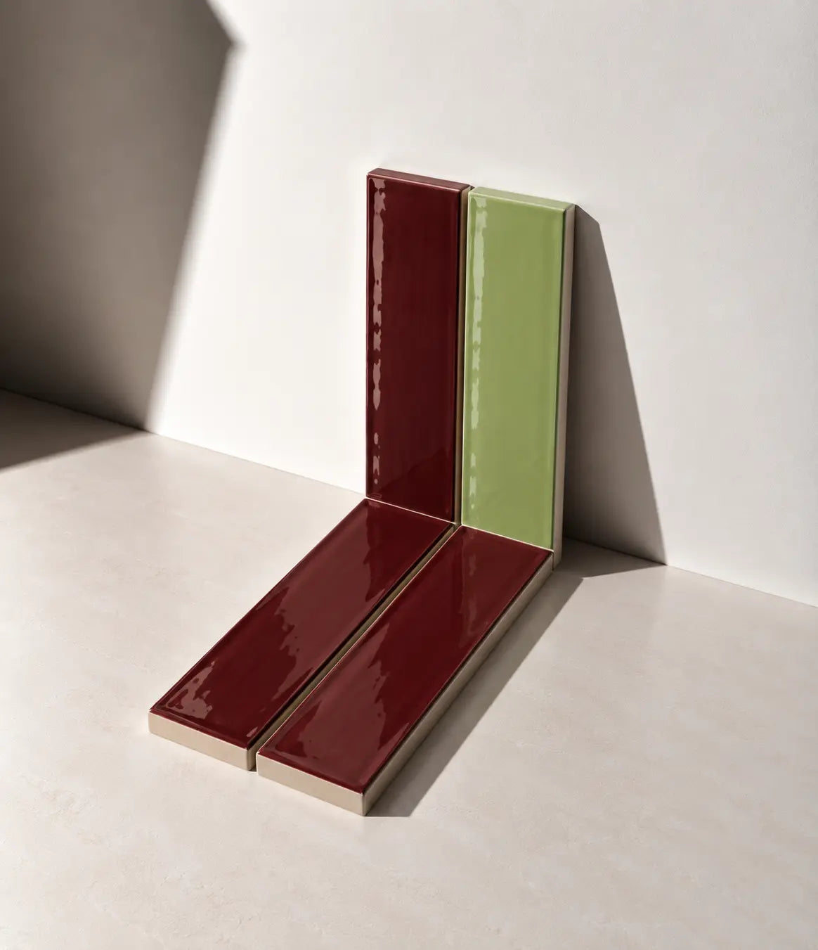 Glossy Burgundy Red and Sage Green handmade ceramic tiles arranged against a white wall in natural sunlight.