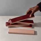 Hand holding a glossy burgundy red rectangular ceramic tile above a stack of handmade tiles on a neutral stone surface.