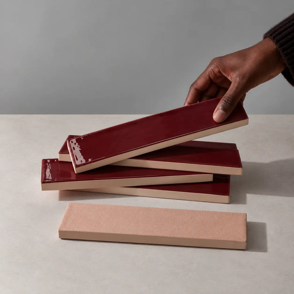 Hand holding a glossy burgundy red rectangular ceramic tile above a stack of handmade tiles on a neutral stone surface.