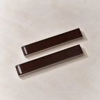 Two glossy dark brown rectangular ceramic tiles with rounded ends, displayed diagonally on a light beige textured background.