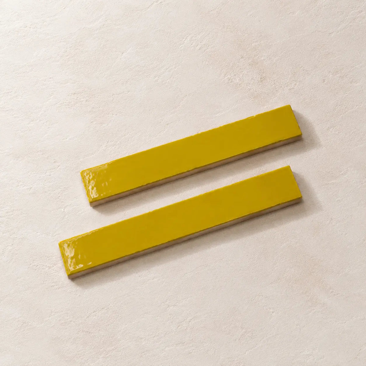 Two glossy citron yellow rectangular ceramic tiles with a textured beige base, arranged diagonally on a light beige textured surface.