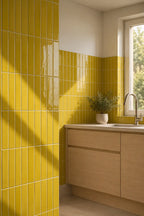 Bright kitchen corner with glossy yellow wall tiles, light wood cabinetry, a white countertop, and a window with natural light.