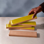 A hand is lifting a glossy citron yellow rectangular tile from a stack of similar tiles, showing the tan back side of the ceramic pieces, set against a neutral gray background.
