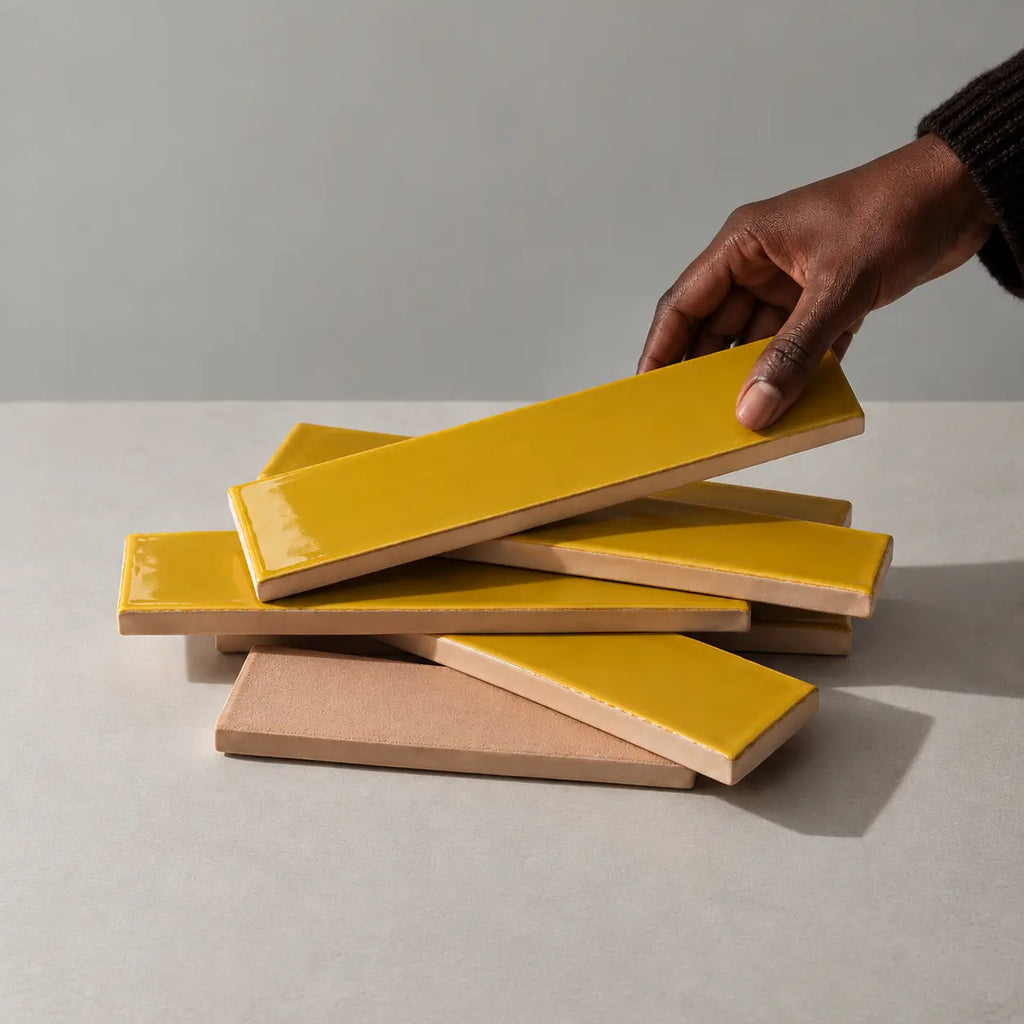 A hand is lifting a glossy citron yellow rectangular tile from a stack of similar tiles, showing the tan back side of the ceramic pieces, set against a neutral gray background.