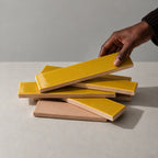 A hand is lifting a glossy citron yellow rectangular tile from a stack of similar tiles, showing the tan back side of the ceramic pieces, set against a neutral gray background.