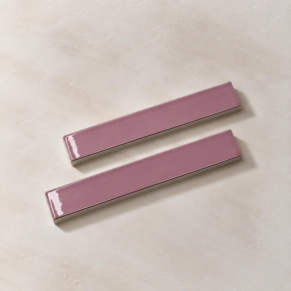 Two glossy rectangular ceramic wall tiles in a dusty mauve-pink color, arranged diagonally on a textured beige background.