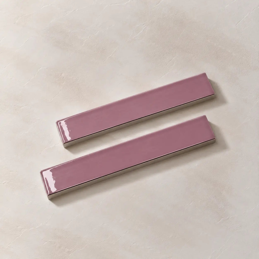 Two glossy rectangular ceramic wall tiles in a dusty mauve-pink color, arranged diagonally on a textured beige background.