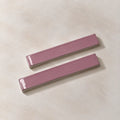 Two glossy rectangular ceramic wall tiles in a dusty mauve-pink color, arranged diagonally on a textured beige background.
