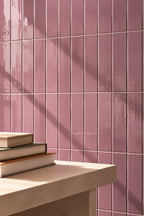 Dusty pink rectangular ceramic wall tiles with a glossy finish, arranged in a grid pattern on a wall above a light beige stone ledge with stacked books.
