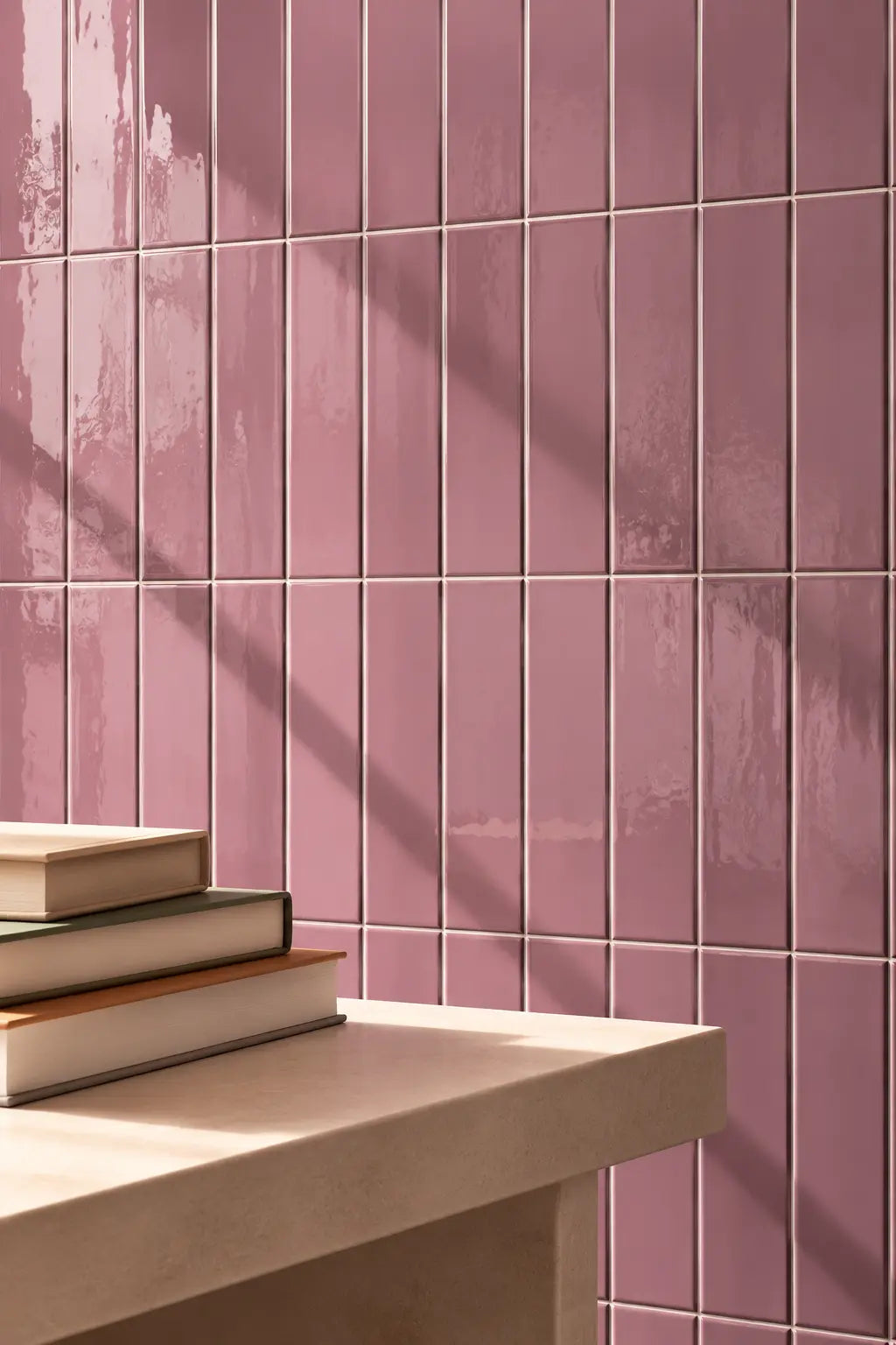 Dusty pink rectangular ceramic wall tiles with a glossy finish, arranged in a grid pattern on a wall above a light beige stone ledge with stacked books.